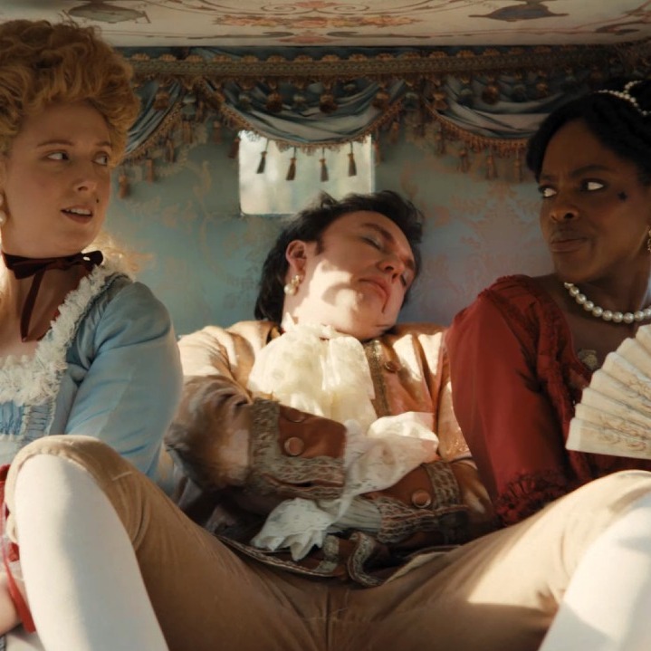 Dos mujeres vestidas de época en un carruaje con un hombre espatarrado y dormido entre ellas