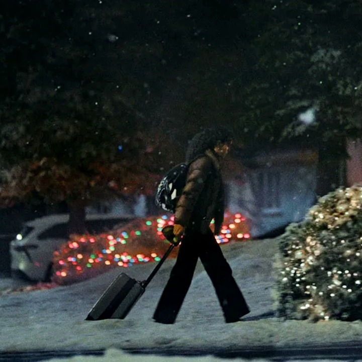 Una chica arrastrando la maleta por la Nieve en la puerta de una casa adornada con luces de Navidad