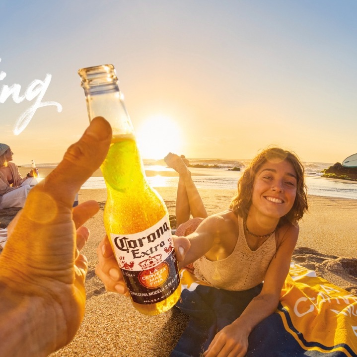Una mano sostiene una Corona y brinda con una chica en un atardecer en la playa