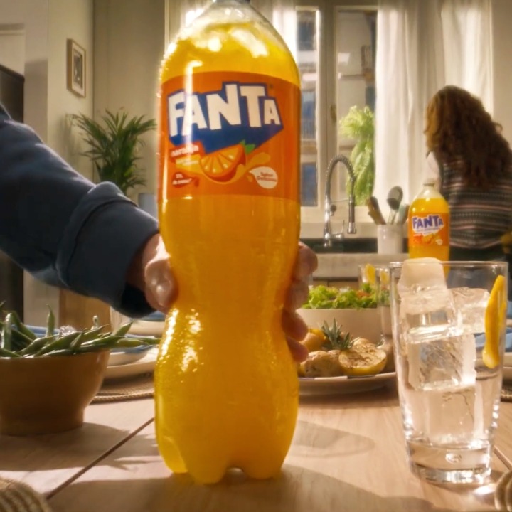 Una mano coloca una botella grande de Fanta sobre una mesa con la comida lista