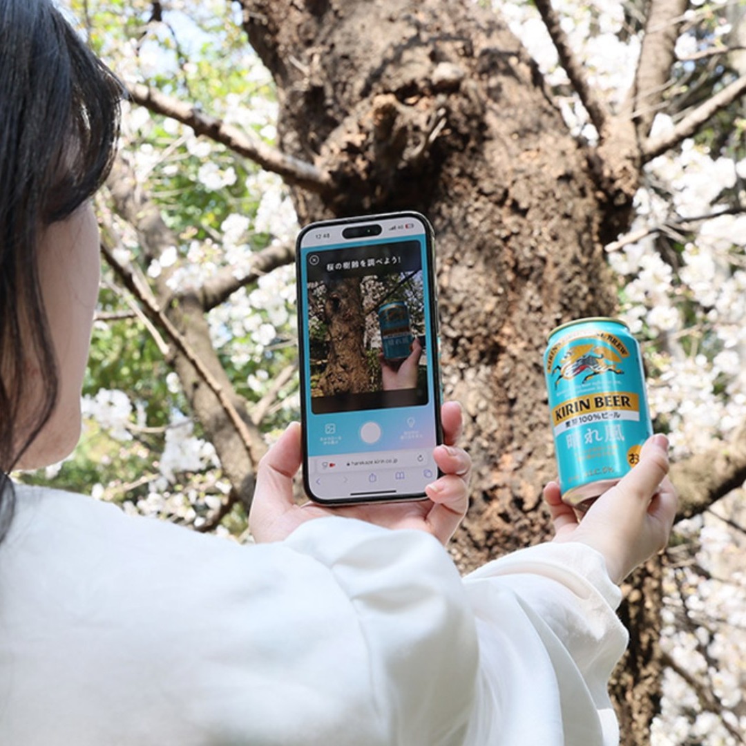 Mujer haciendo una foto a una lata de Kirin con el móvil, con un cerezo en flor de fondo
