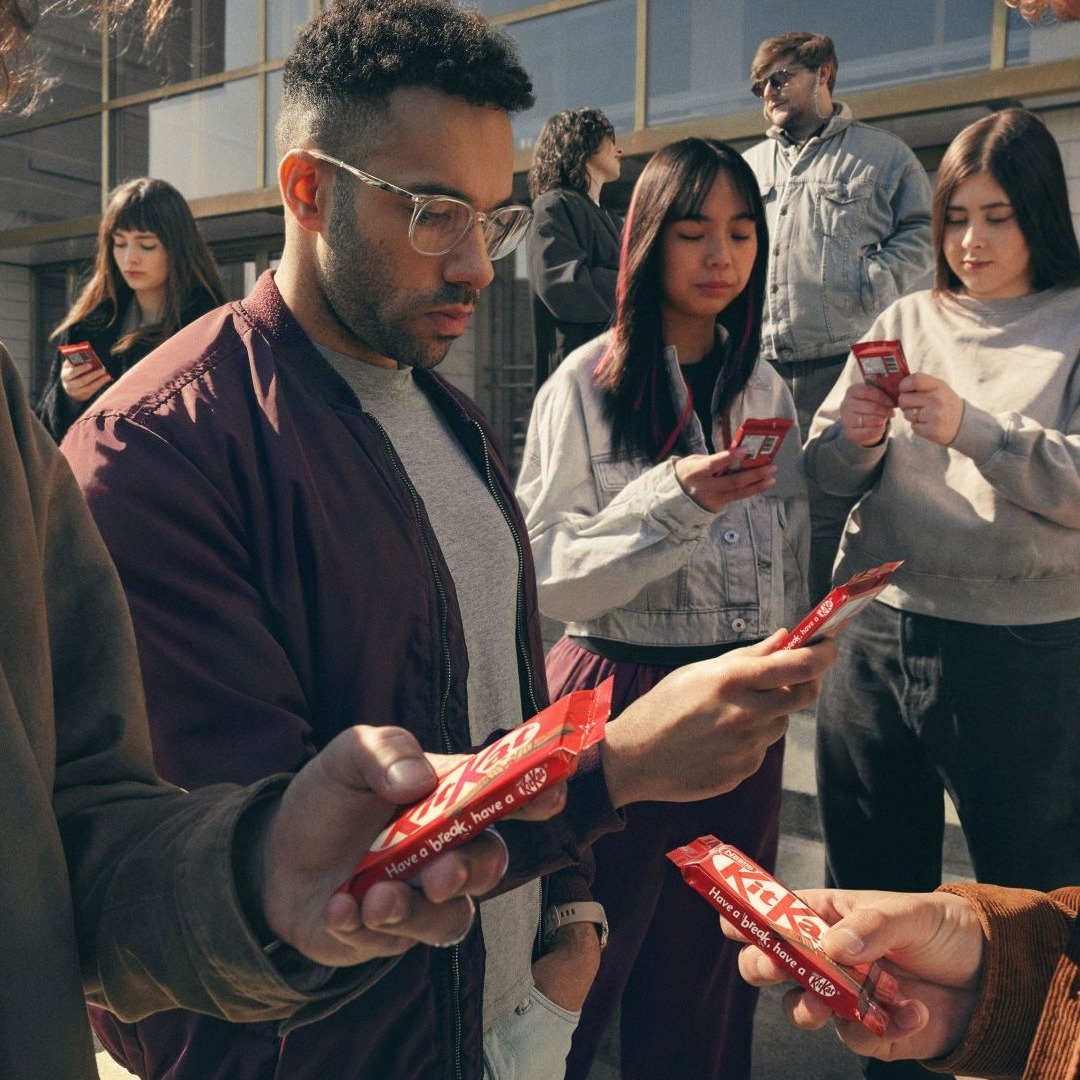 Personas mirando sus teléfonos móviles en la calle, pero en lugar de móviles sujetan kitkats con sus manos