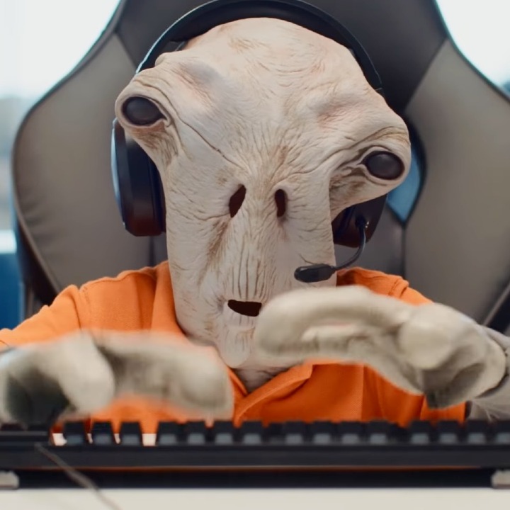 un marciano vestido con el uniforme de pccomponentes sentado delante del ordenador con unos auriculares de centralita