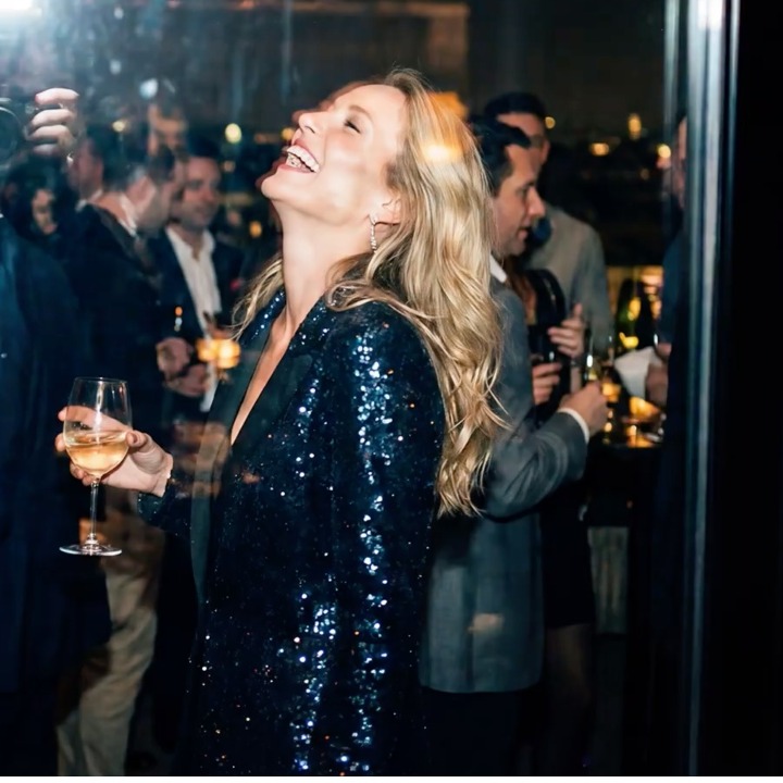 Una mujer rubia con vestido de lentejuelas riendo en una fiesta con una copa de vino