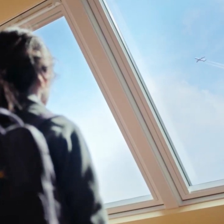 Una niña mirando por la ventada de Velux viendo un avión volar en el cielo