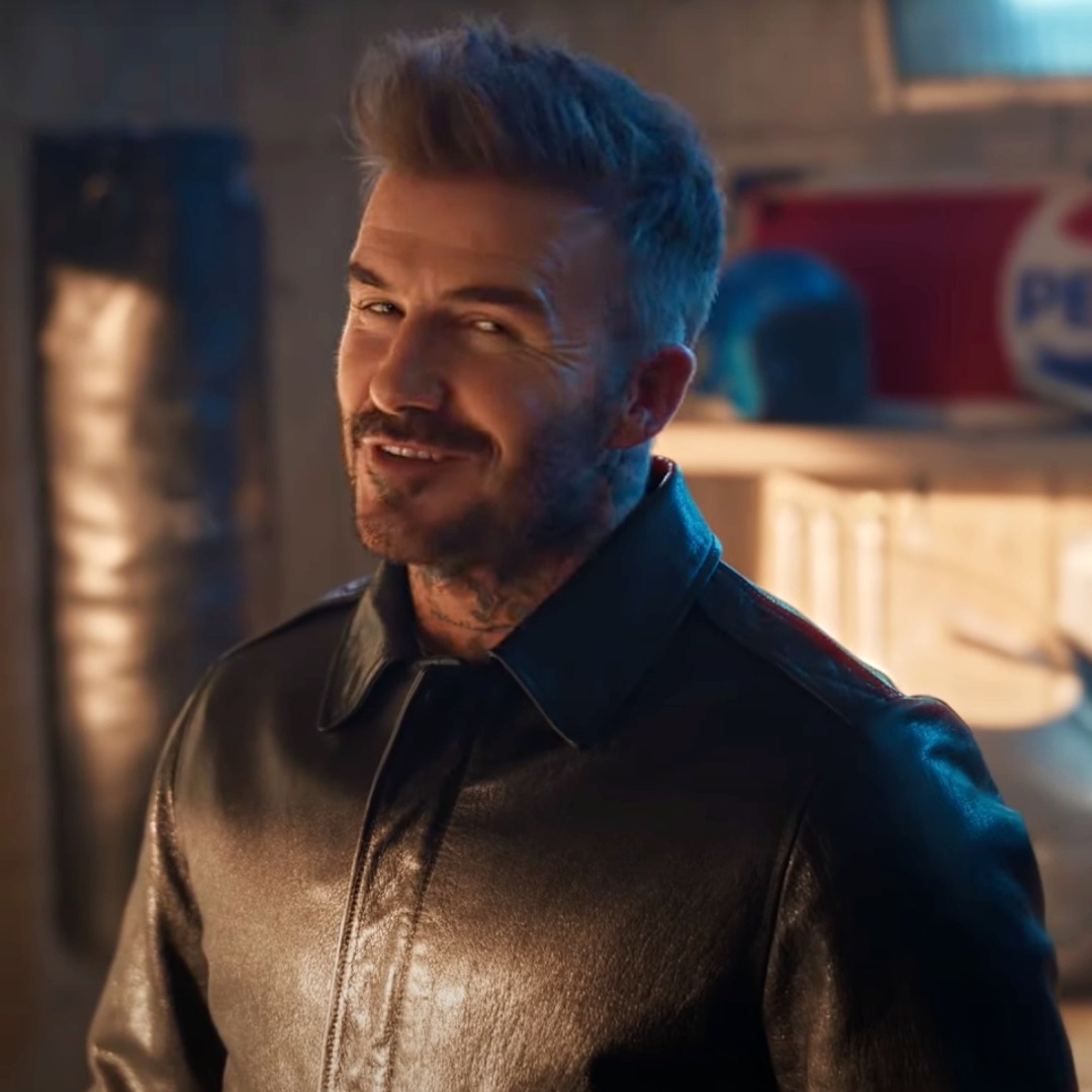 David Beckham sonriendo a cámara desde un garaje con un cartel de pepsi al fondo.