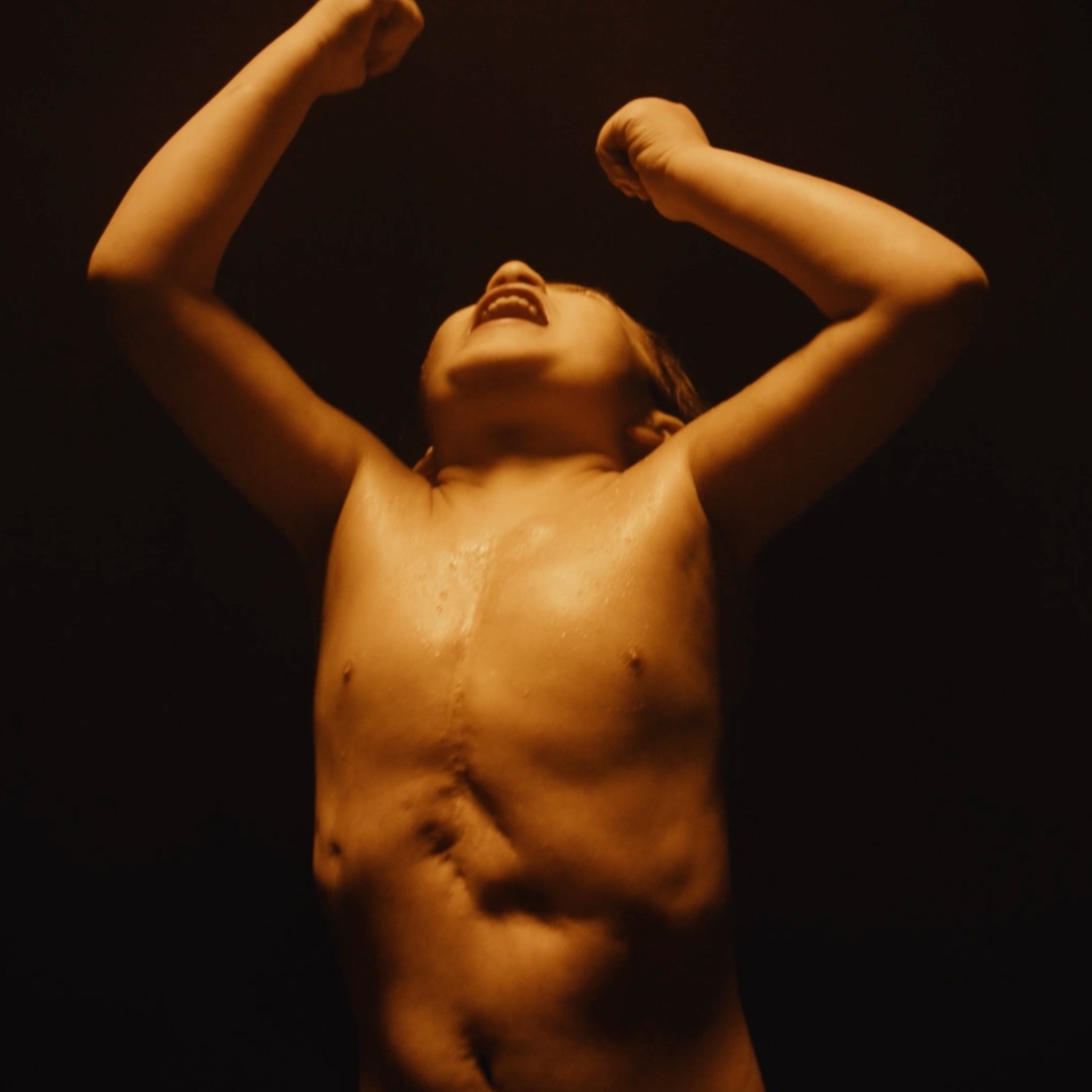 Niño sin camiseta gritando con los brazos en alto sobre un fondo negro