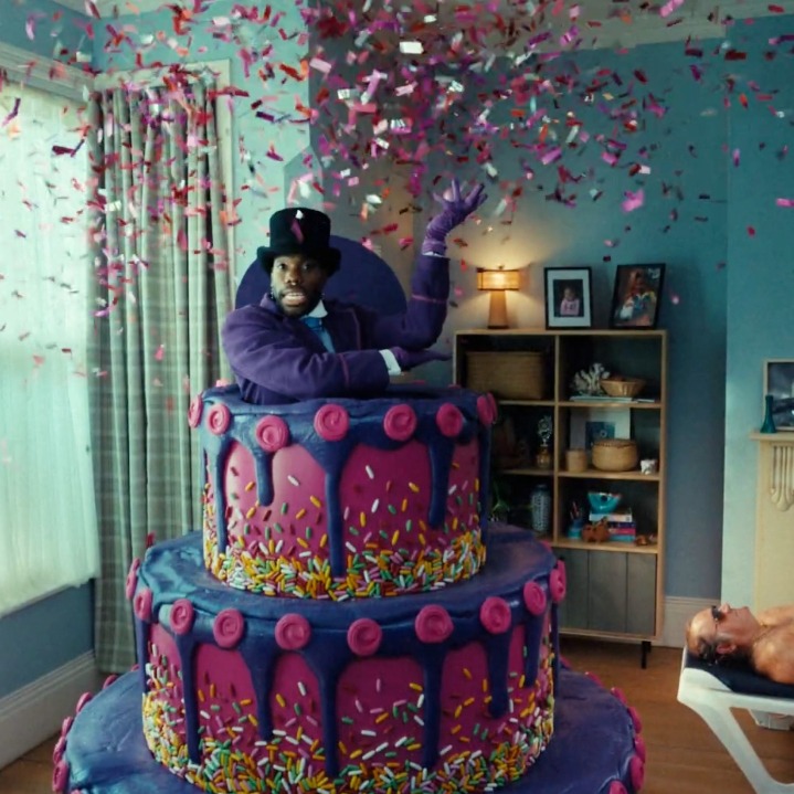 Un hombre saliendo de una tarta morada y rodeado de confeti en medio de un salón de casa
