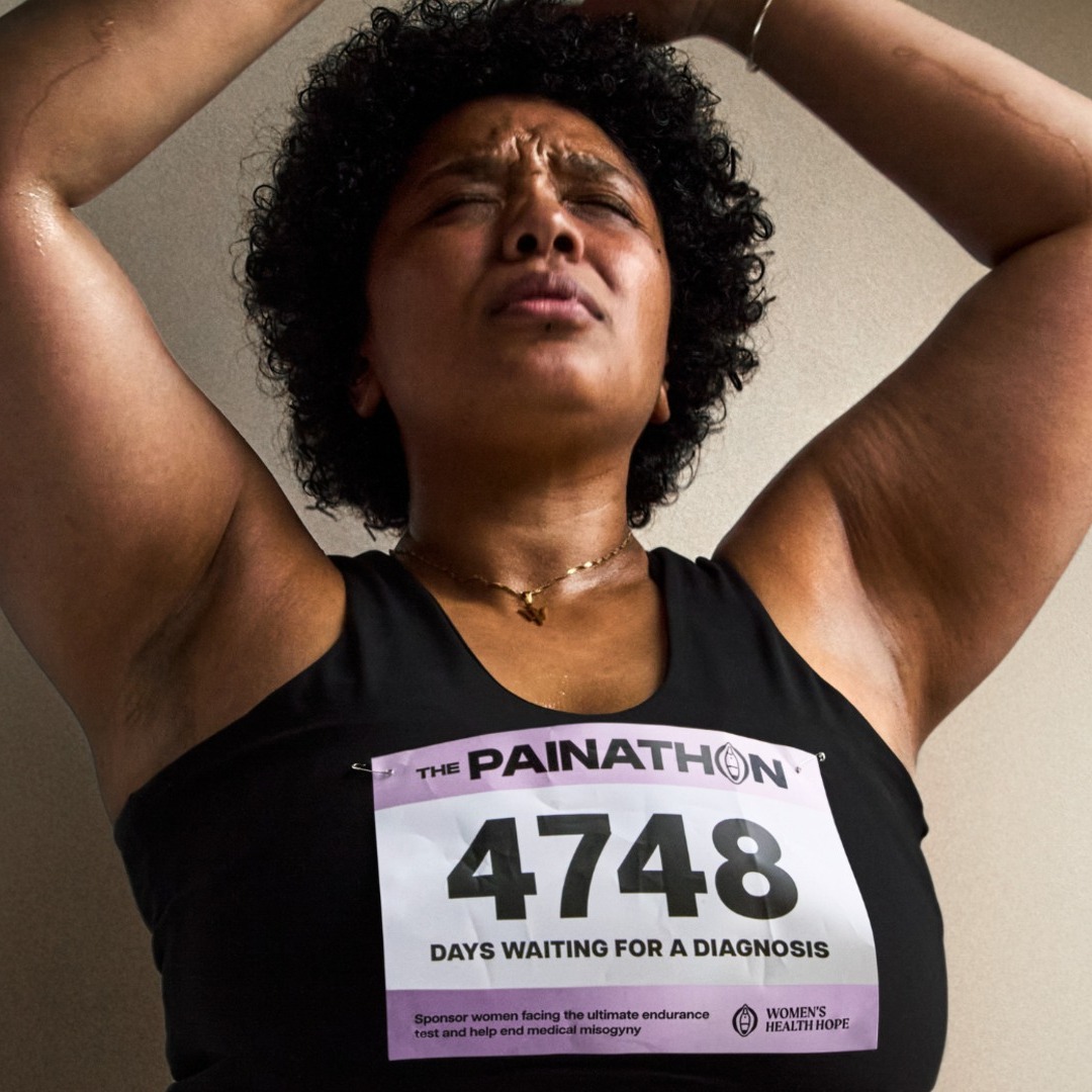 Mujer con expresión de dolor levantando los brazos con los ojos cerrados, y un dorsal de la campaña Painathon en la camiseta.