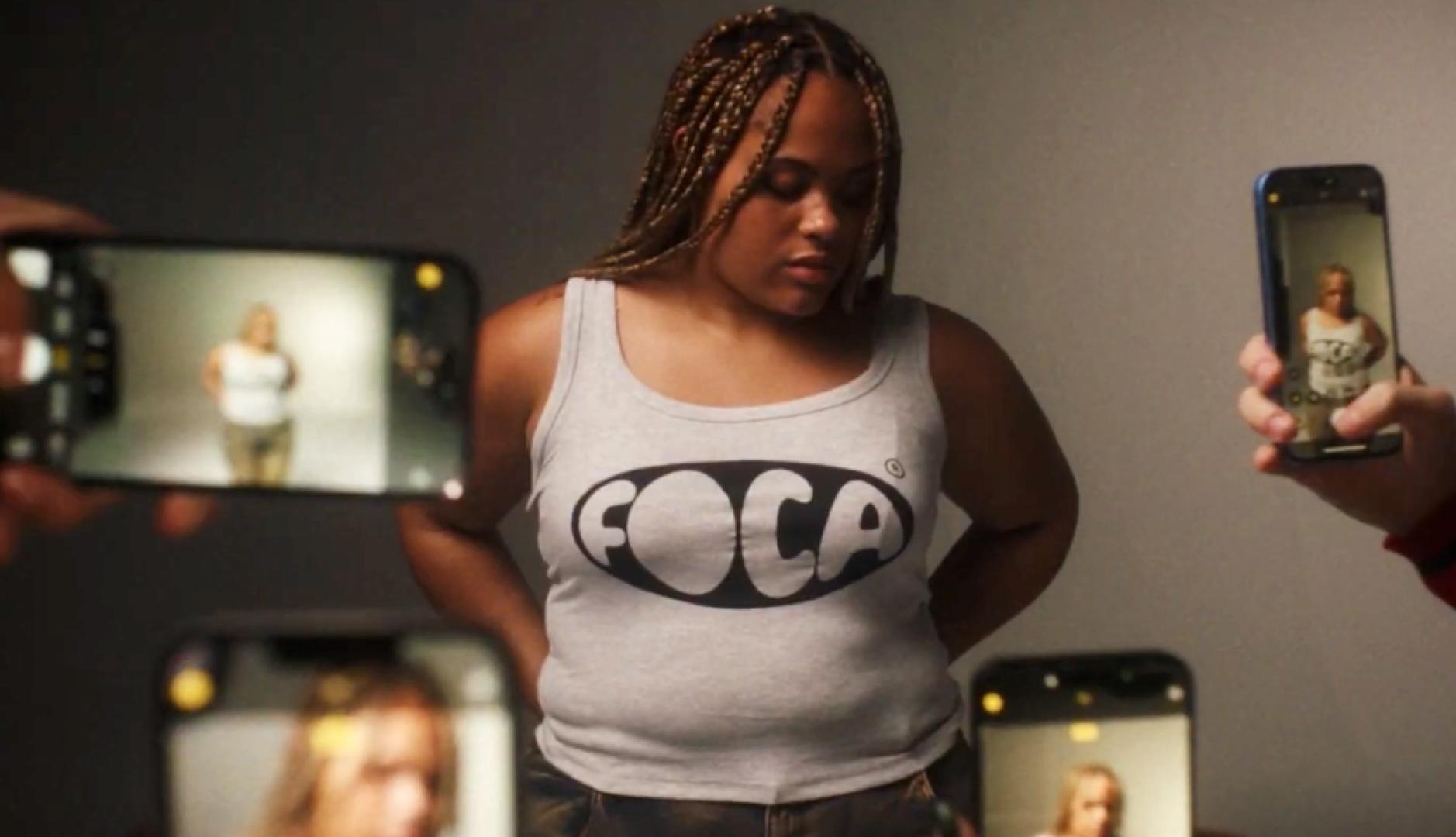 Una chica lleva una camiseta que reza "foca" muentras varias manos con móviles le hacen fotos