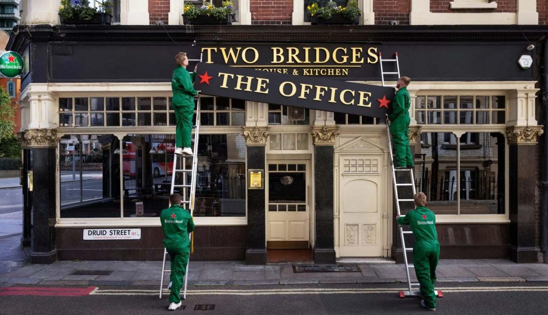 Heineken celebra el trabajo presencial convirtiendo los pubs en extensiones de la oficina Trabajadores con monos de Heineken colocan un cartel en la fachada de un pub