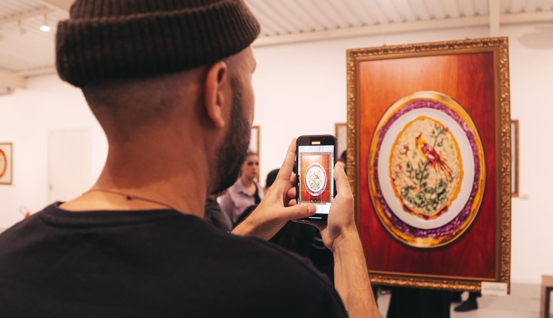 Persona joven de espaldas haciendo una foto con su móvil a uno de los cuadros de ensalada de patatas, de la exposición "Food Art", integrada en la campaña "Mayonismo" de Hellmann's Brasil