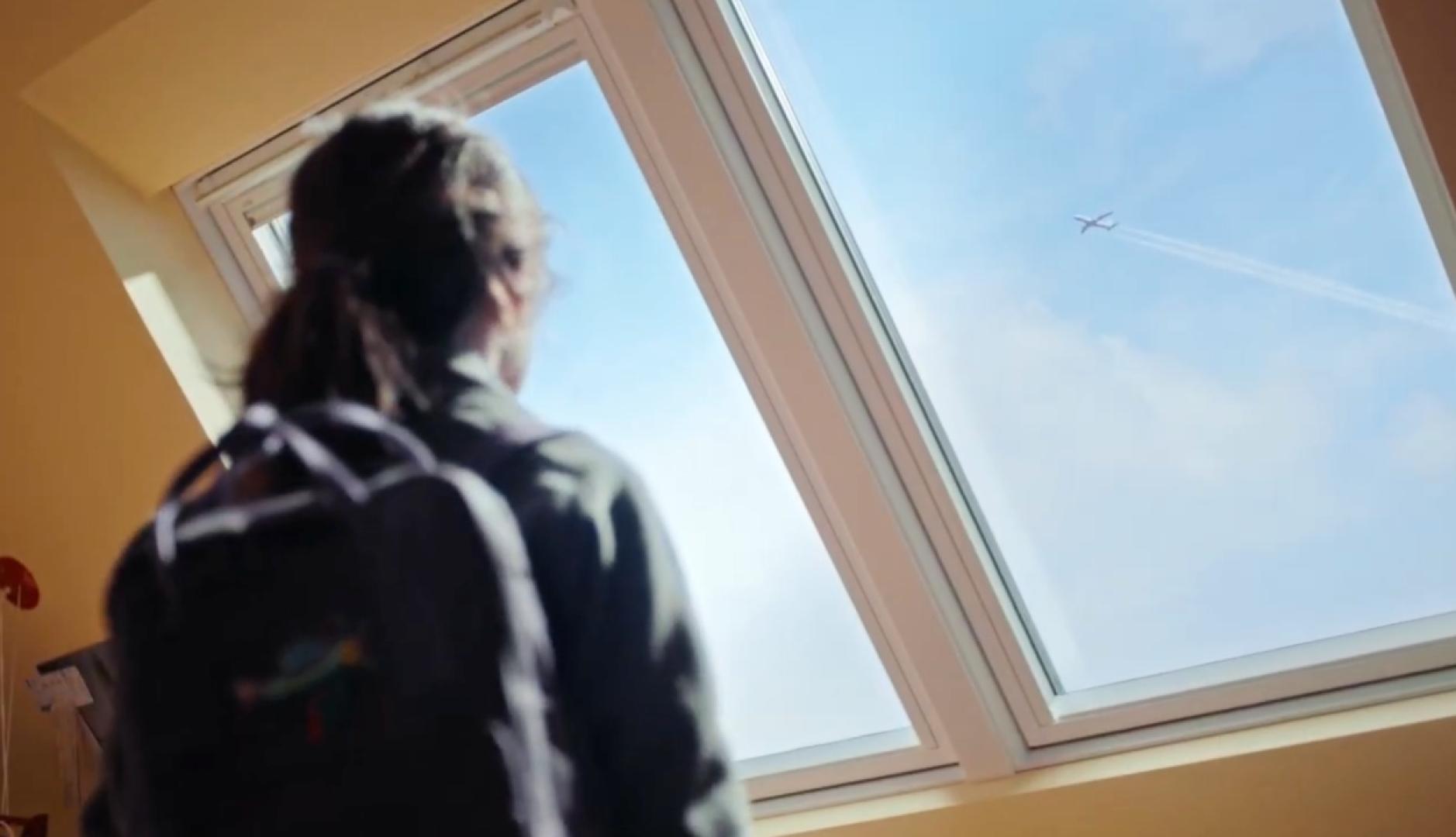 Una niña mirando por la ventada de Velux viendo un avión volar en el cielo