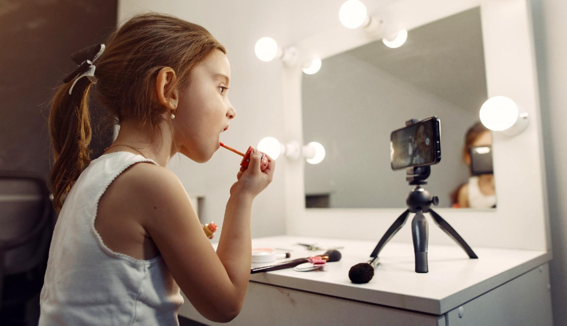 Una niña pequeña maquillándose en un espejo con un teléfono que graba