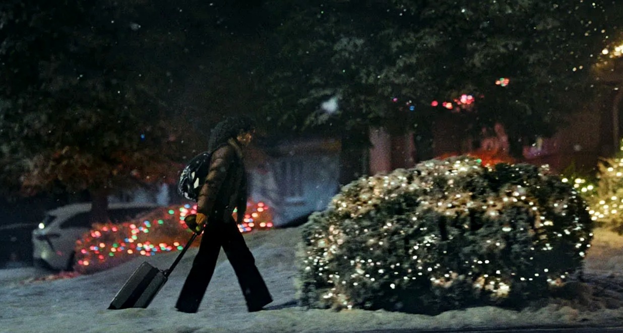 Una chica arrastrando la maleta por la Nieve en la puerta de una casa adornada con luces de Navidad