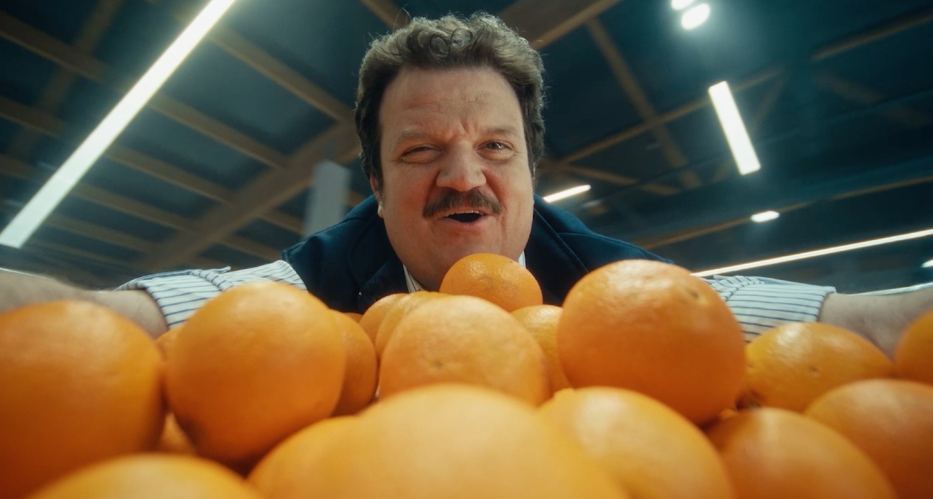 Un cajero canta apoyándose en la estantería de las naranjas de un supermercado