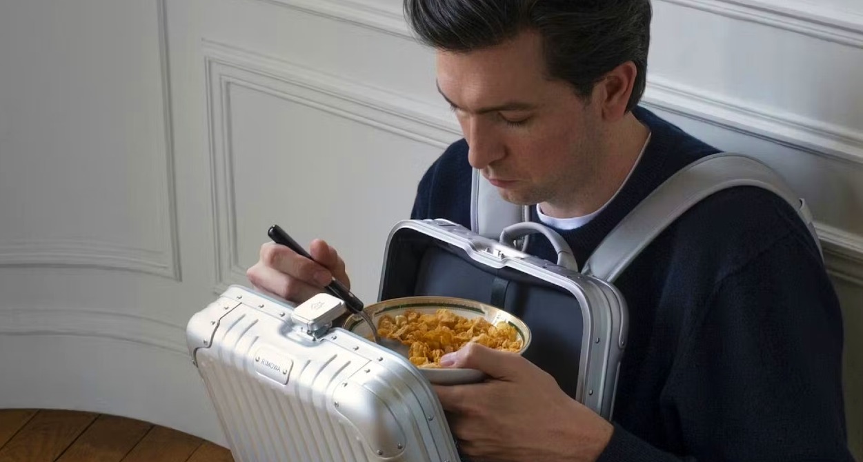 Nicholas Braun sentado en el suelo comiendo cereales dentro de la mochila de Rimowa