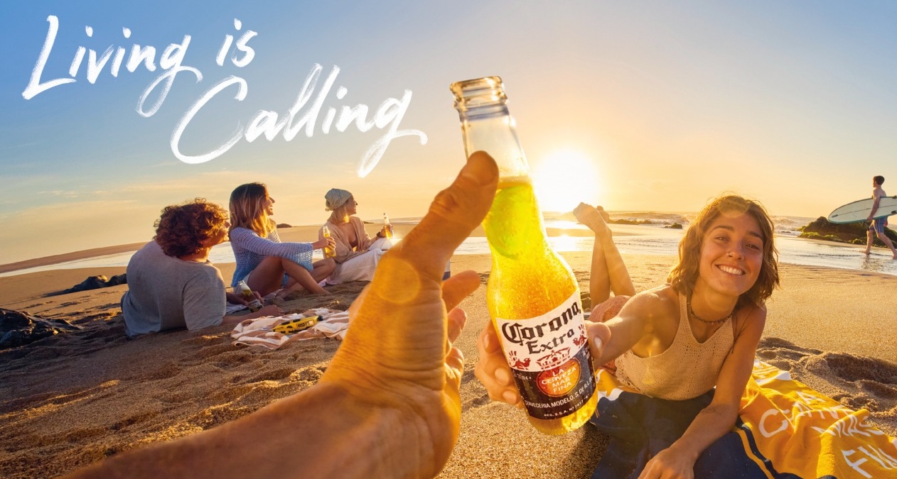 Una mano sostiene una Corona y brinda con una chica en un atardecer en la playa
