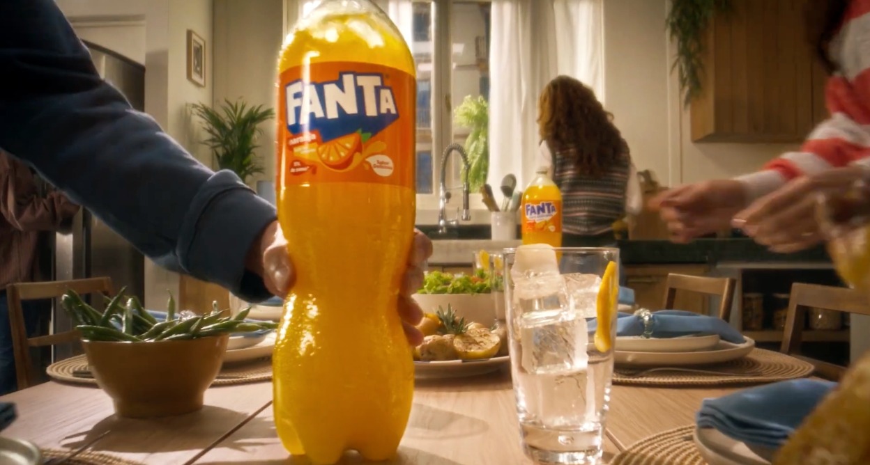 Una mano coloca una botella grande de Fanta sobre una mesa con la comida lista