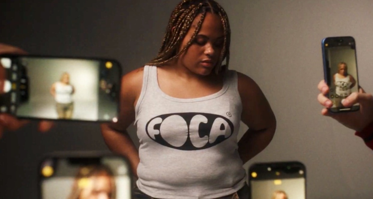 Una chica lleva una camiseta que reza "foca" muentras varias manos con móviles le hacen fotos