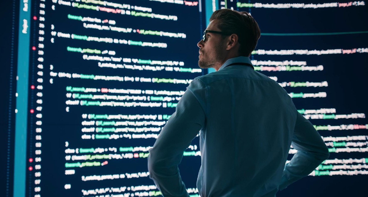 Un hombre con los brazos en jarras mira unas pantallas grandes con código HTML
