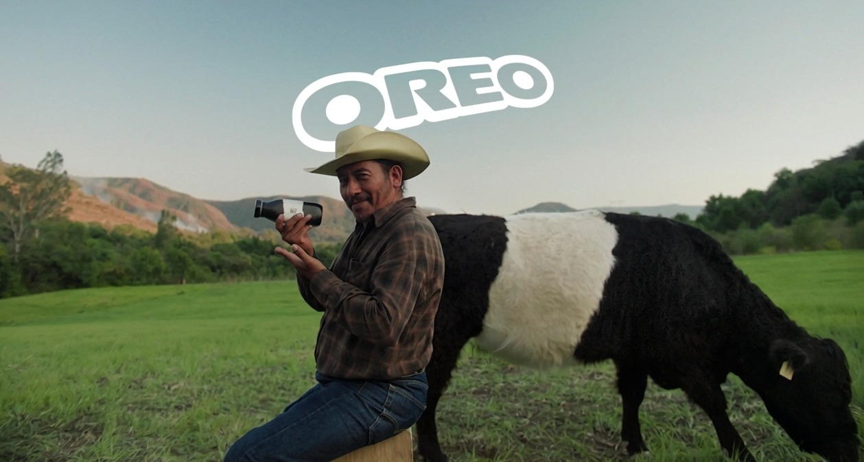 Un granjero posando con la botella de leche de vaca Oreo y detrás una vaca Belted Galloway