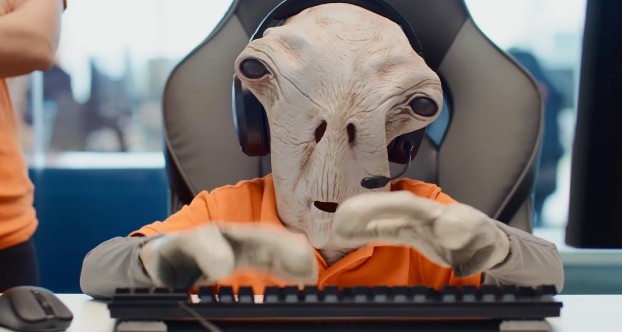un marciano vestido con el uniforme de pccomponentes sentado delante del ordenador con unos auriculares de centralita