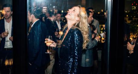 Una mujer rubia con vestido de lentejuelas riendo en una fiesta con una copa de vino