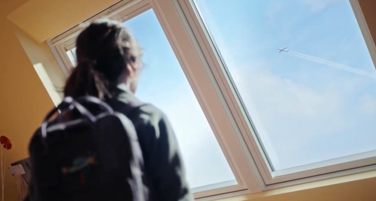 Una niña mirando por la ventada de Velux viendo un avión volar en el cielo