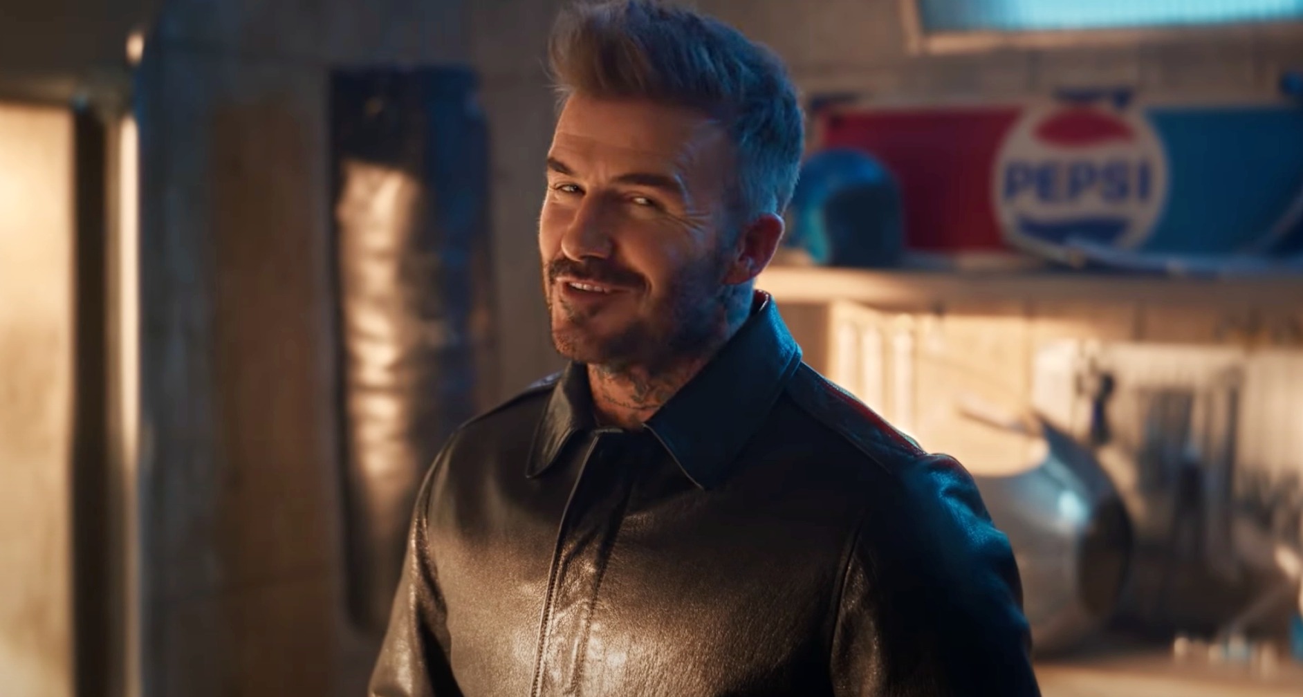 David Beckham sonriendo a cámara desde un garaje con un cartel de pepsi al fondo.
