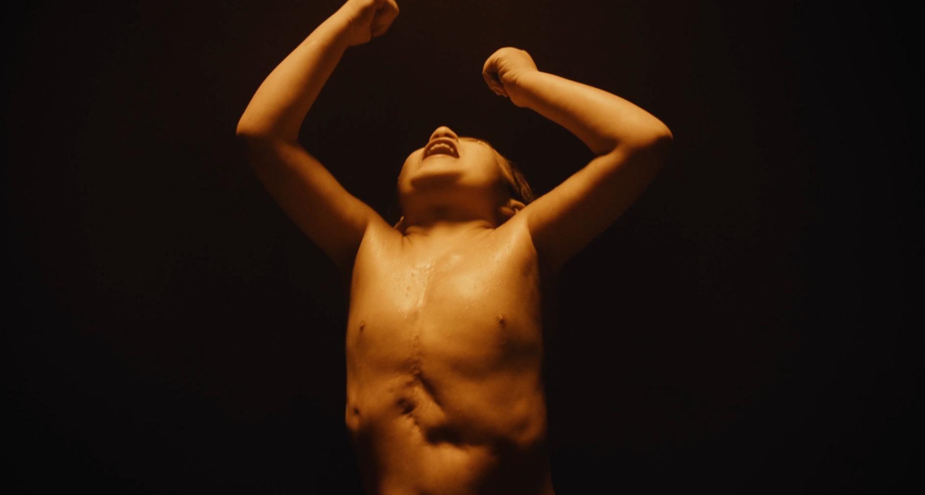 Niño sin camiseta gritando con los brazos en alto sobre un fondo negro