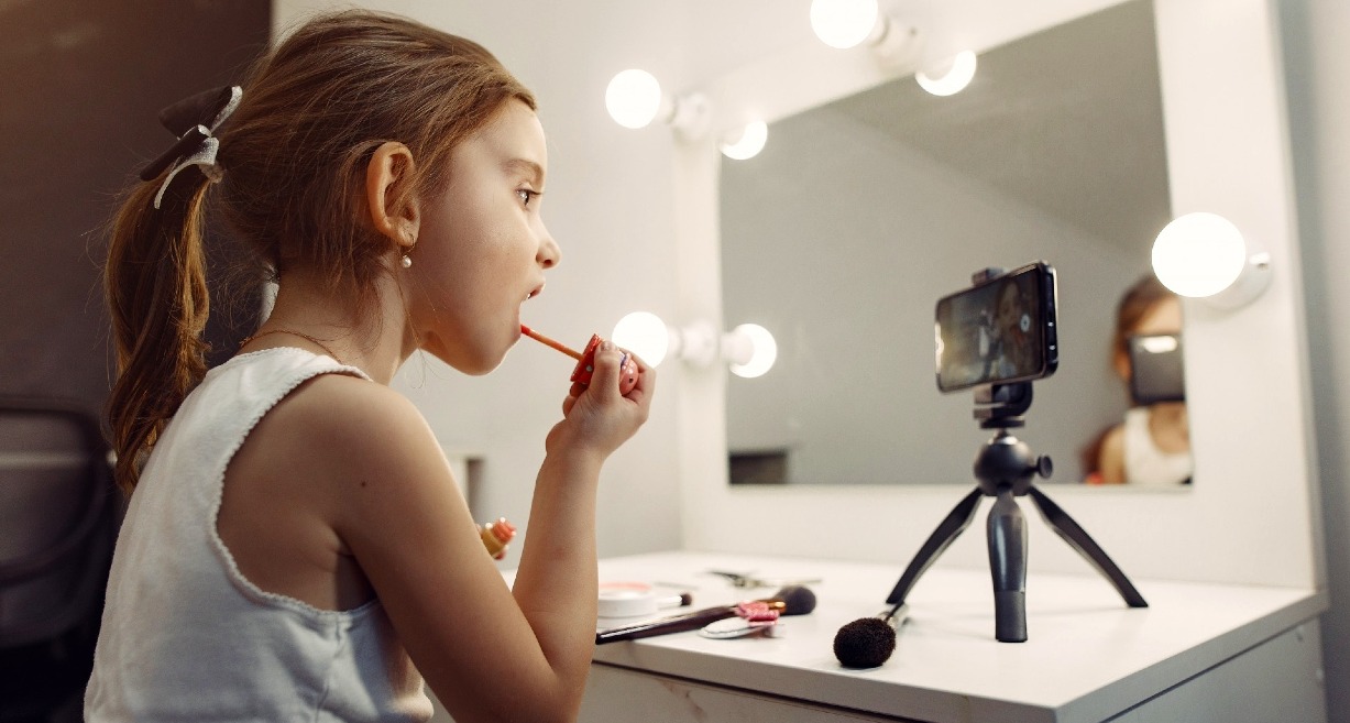 Una niña pequeña maquillándose en un espejo con un teléfono que graba