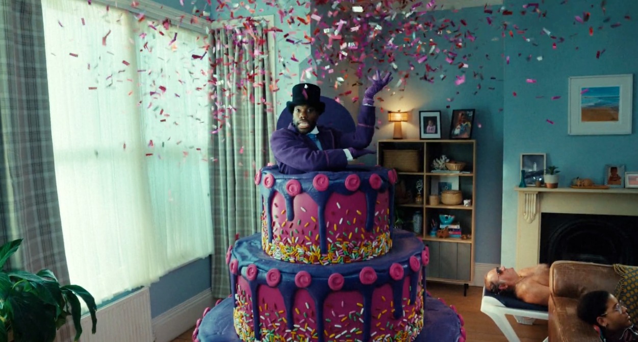 Un hombre saliendo de una tarta morada y rodeado de confeti en medio de un salón de casa