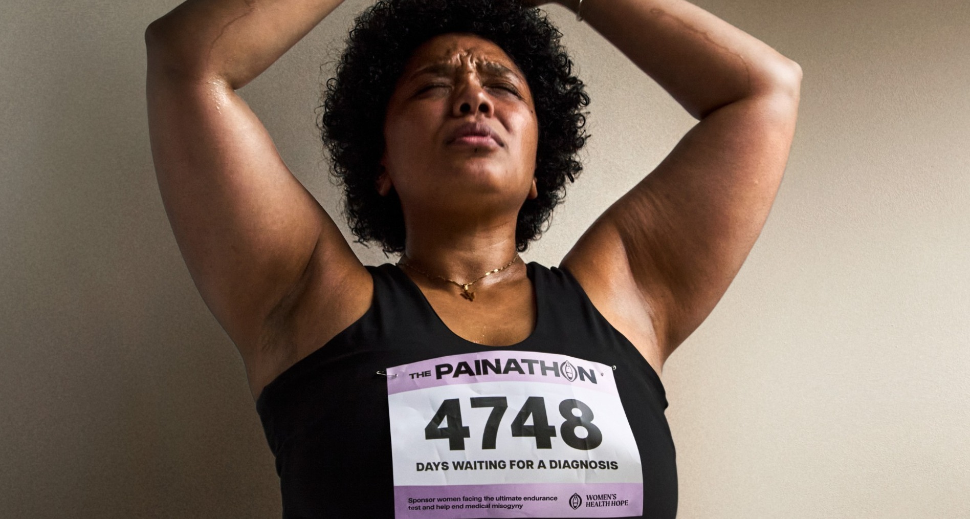 Mujer con expresión de dolor levantando los brazos con los ojos cerrados, y un dorsal de la campaña Painathon en la camiseta.