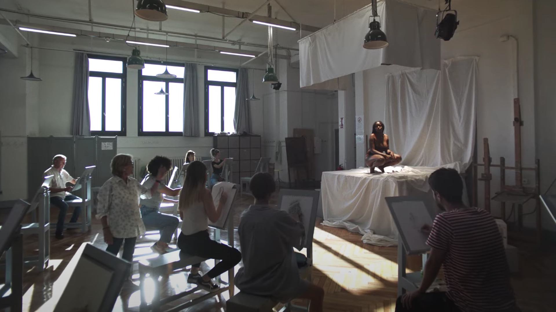 Imagen del spot: una chica posando como modelo, sentada en cuclillas, en una clase de dibujo.